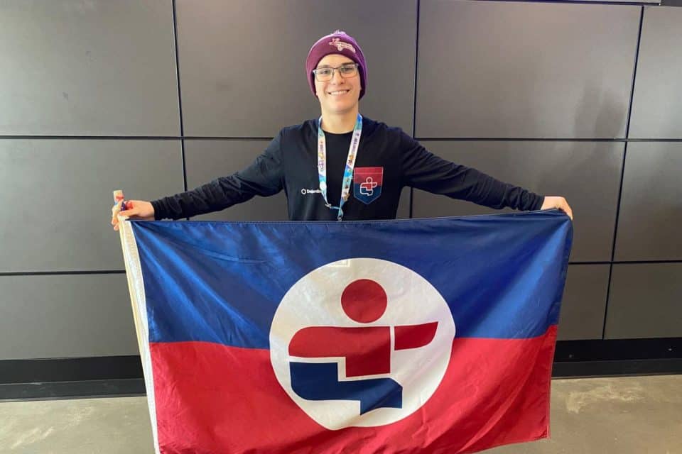 Intérieur sobre avec mur gris; une personne tient un drapeau coloré et porte une médaille autour du cou.