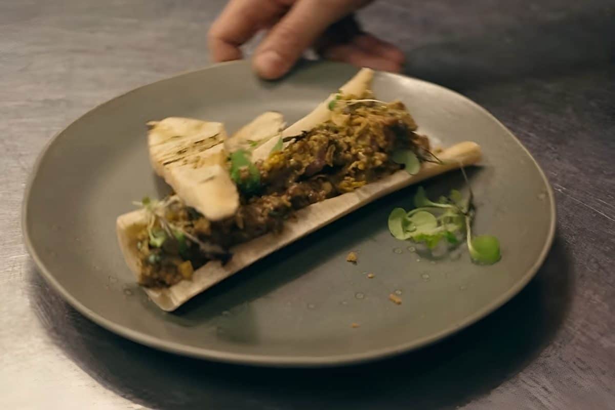 Assiette grise sur une table sombre avec un os garni de viande et d’herbes, accompagné de morceaux grillés et de pousses vertes.