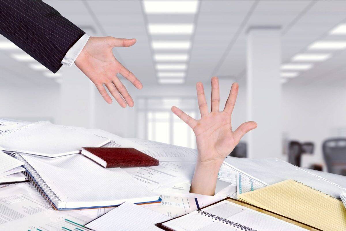Pile de dossiers et cahiers sur un bureau dans un bureau lumineux; une main sort des papiers tandis qu’une autre se tend au-dessus pour aider.