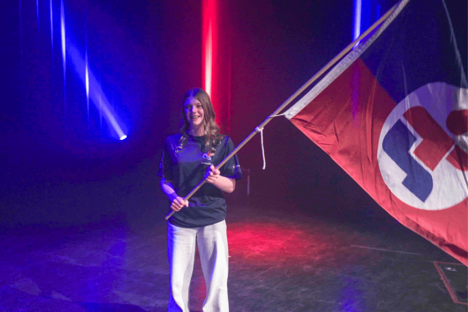 Sur une scène sombre éclairée de faisceaux bleus et rouges, Emma Larson tient le drapeau de Launaudière.