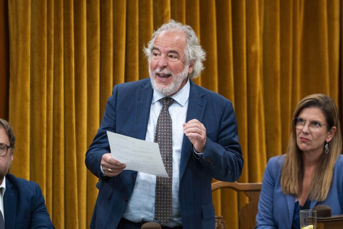 Dans une salle parlementaire aux murs boisés, Rhéal Fortin debout lit un document, entouré de collègues assis, microphones et pupitres visibles.