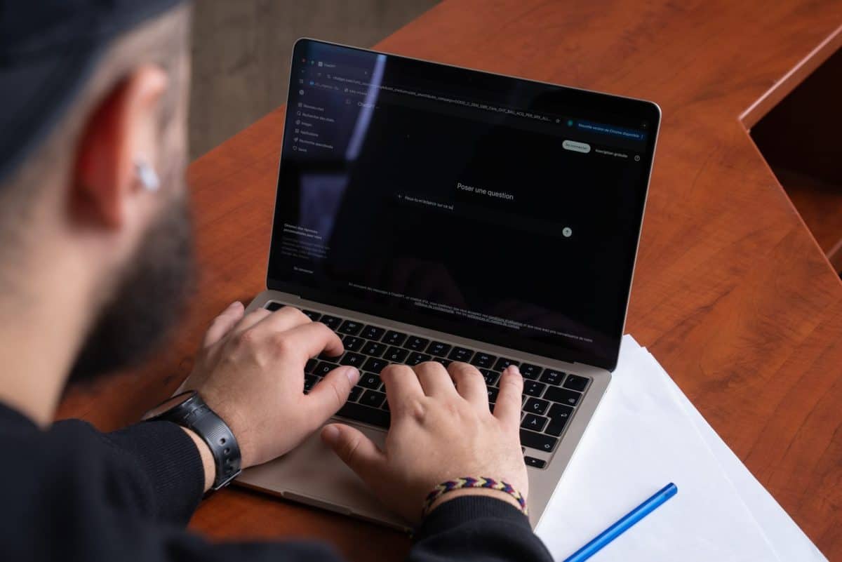 Ordinateur portable sur table en bois, écran ouvert sur une interface de texte, avec carnet et stylo à côté.