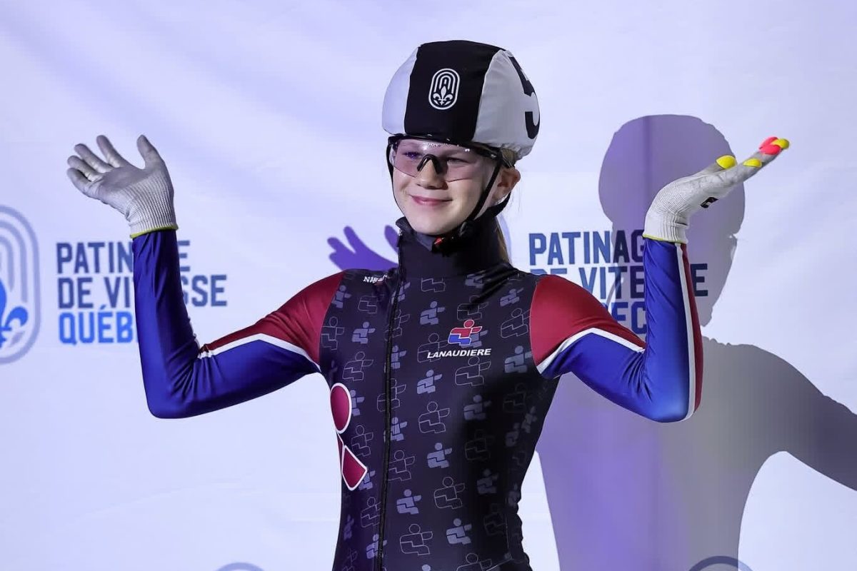 Maïka Scholten en combinaison aux couleurs de Lanaudière pose devant un mur « Patinage de vitesse Québec », casque et lunettes en place.