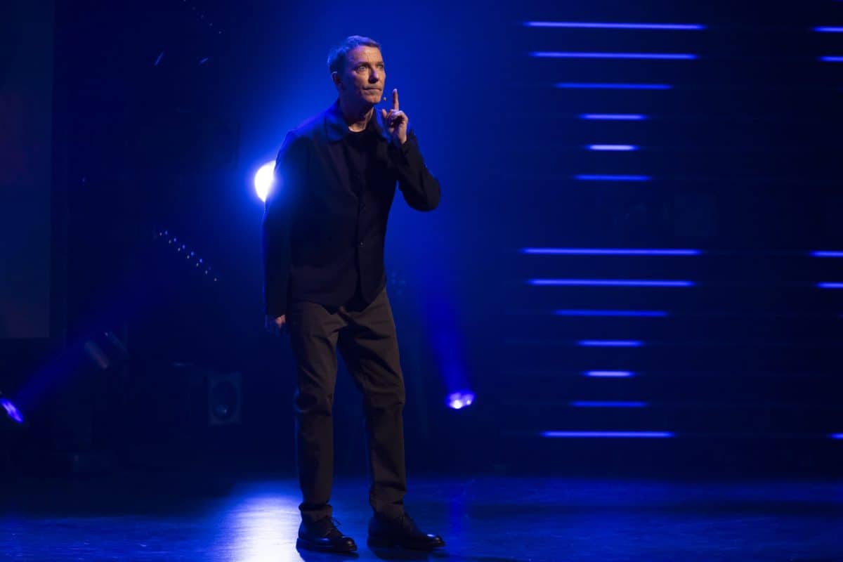 Scène sombre éclairée en bleu; Patrice L’Écuyer sur scène, sous projecteurs, dans une ambiance de spectacle.