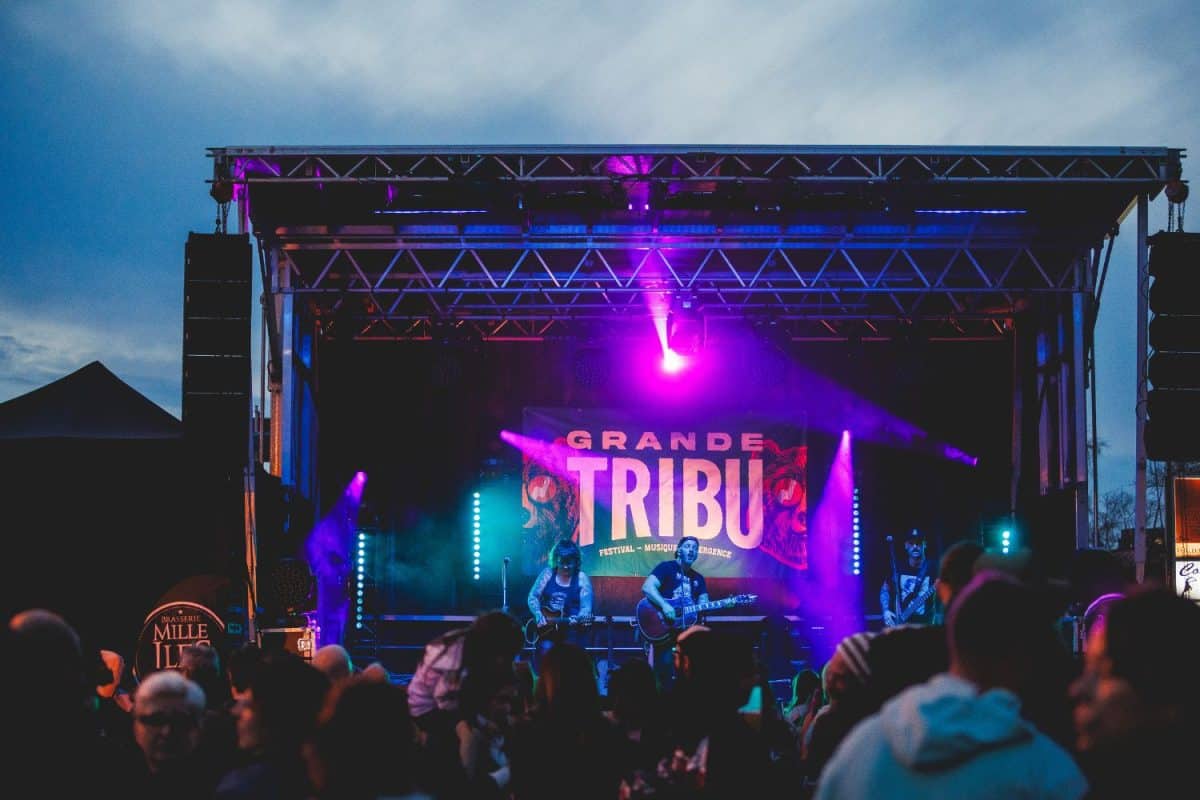 Groupe musical en concert sur scène avec éclairage violet et bleu devant foule sous ciel dégagé