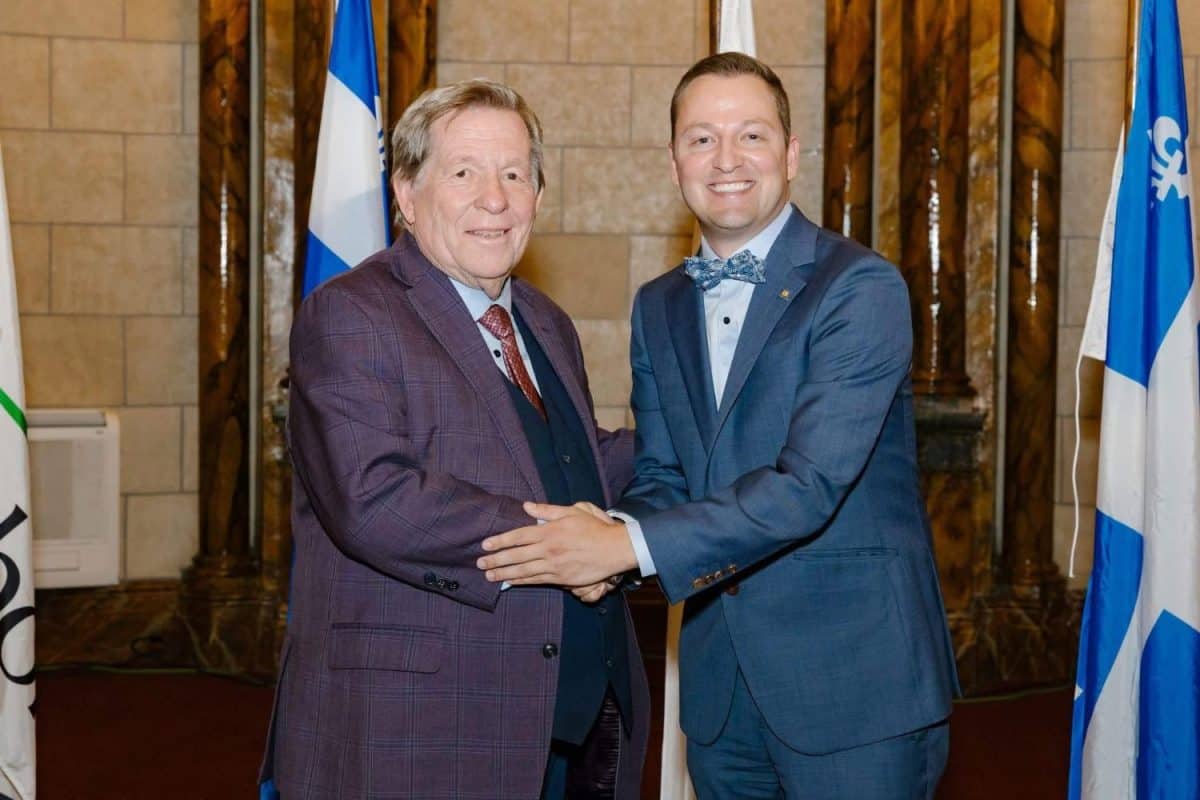 Deux hommes en costume sombre se serrant la main devant drapeaux québécois et canadiens dans un édifice