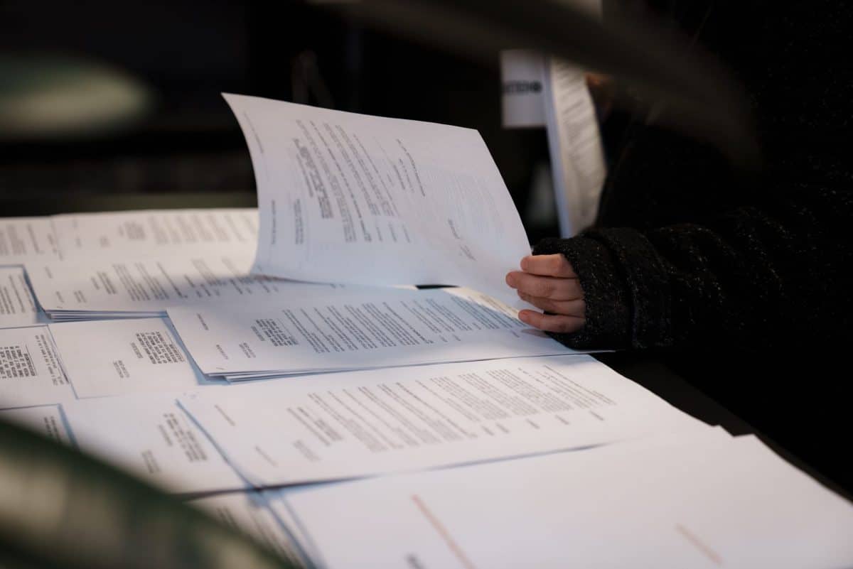 Table couverte de documents imprimés, pages manipulées à la main dans un environnement intérieur sombre, ambiance de consultation administrative.