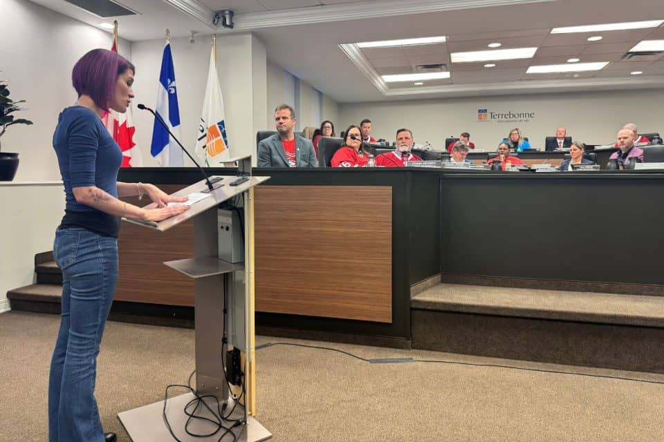 Citoyenne prenant la parole au podium lors d'un conseil municipal de Terrebonne, élus assis au banc devant les drapeaux