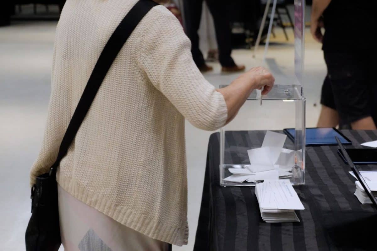Personne déposant un bulletin dans une boîte transparente sur une table, dans un lieu intérieur servant de bureau de vote.