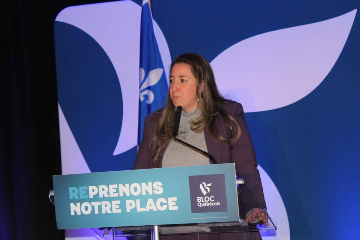 Une scène intérieure avec pupitre et micro devant un fond bleu aux symboles du Bloc québécois et un drapeau du Québec à l’arrière-plan.