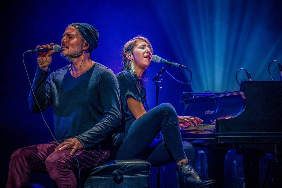 Deux artistes sur scène éclairée en bleu, assis dos à dos près d’un piano, chantant chacun dans un micro lors d’une performance musicale.