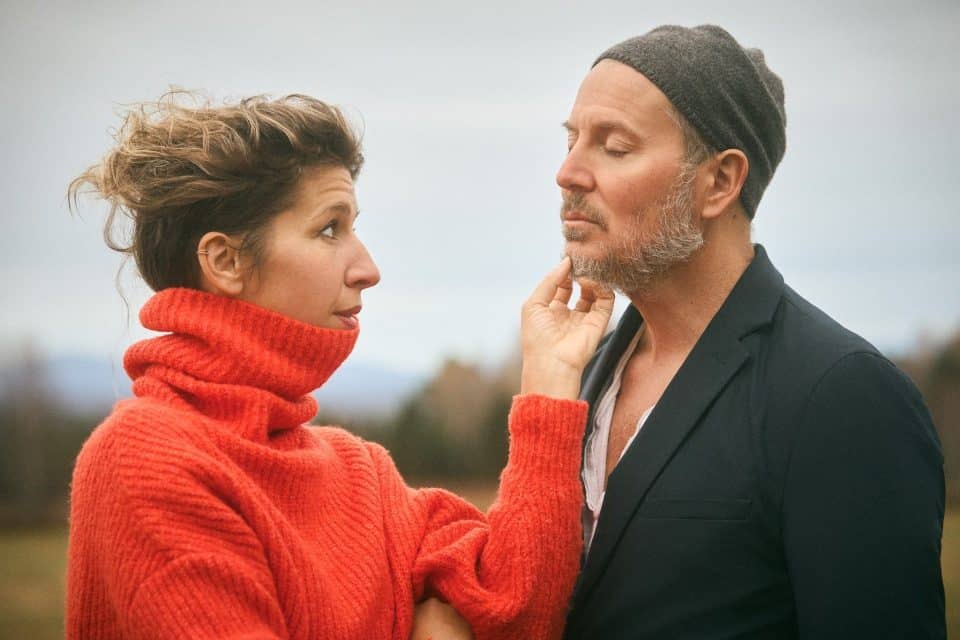 Deux personnes se font face en extérieur, dans un paysage flou, l’une touchant le menton de l’autre dans un moment calme et rapproché.