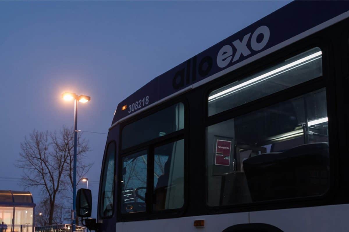 Autobus exo stationné près d’un abribus éclairé au crépuscule, avec lampadaires allumés, route mouillée et arbres sans feuilles en arrière-plan.