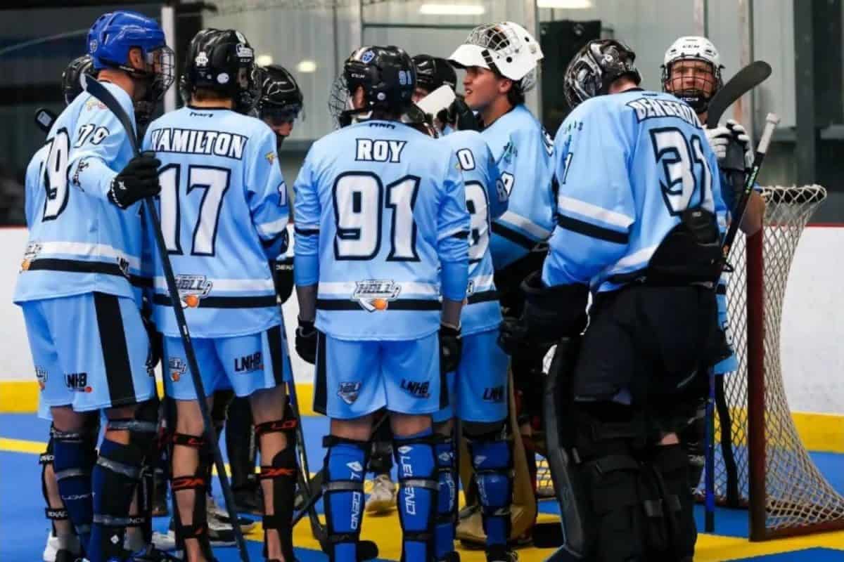 Aréna intérieur avec surface bleue, filet de hockey et joueurs regroupés près du but, ambiance sportive avec bandes et vitre autour de la patinoire.