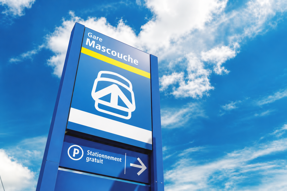 Panneau de la gare Mascouche avec logo de train, dressé sur fond de ciel bleu parsemé de nuages blancs