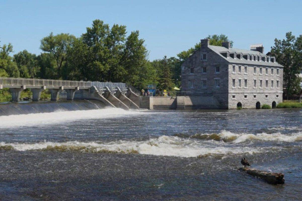 Barrage déversant ses eaux dans la rivière des Mille Îles sous un ciel bleu, moulin historique en pierre sur la rive