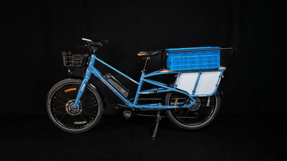 Vélo cargo électrique bleu équipé d’un panier avant et d’un grand compartiment de transport arrière, présenté sur fond noir en studio.