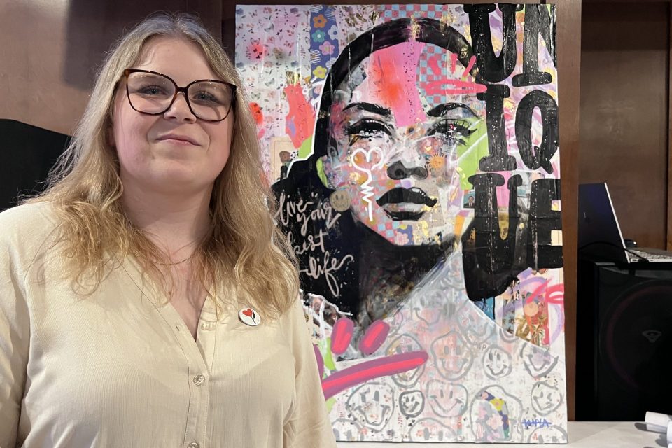 Personne posant à l’intérieur devant une grande toile colorée mêlant collage, portrait stylisé et motifs graphiques, lors d’une exposition artistique.