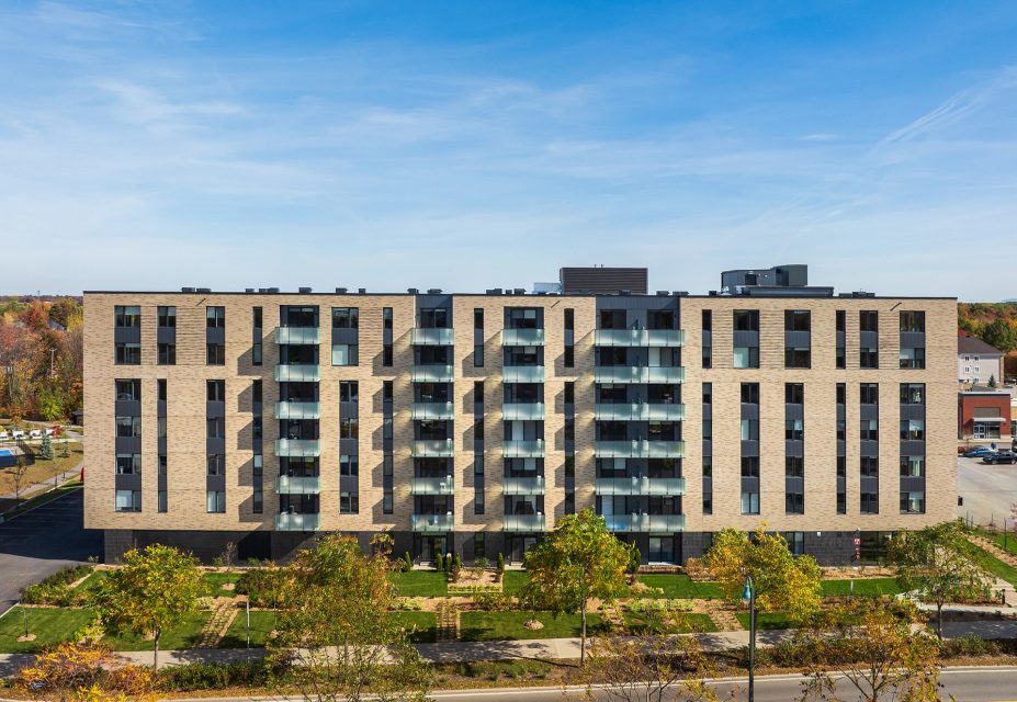 Immeuble résidentiel en brique beige avec balcons vitrés, entouré d'arbres aux couleurs automnales sous un ciel bleu