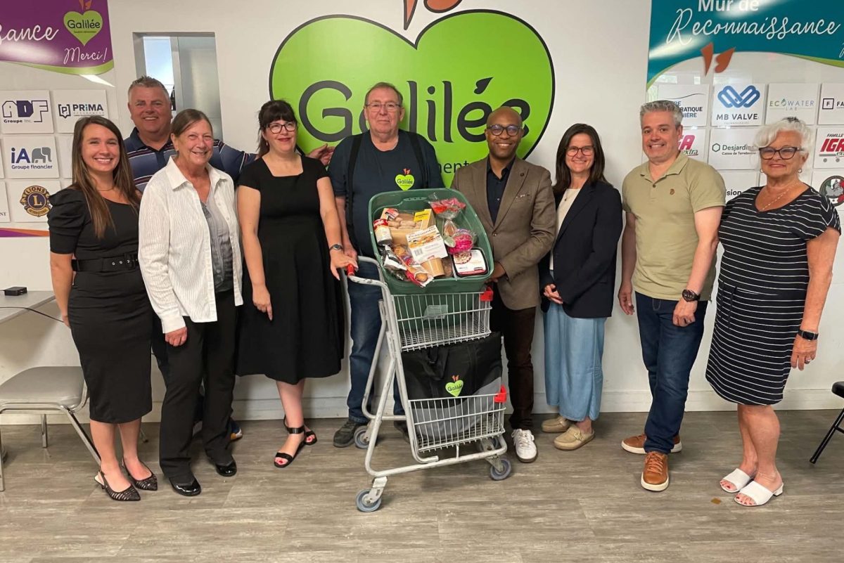 Groupe de personnes posant autour d'un panier d'épicerie rempli devant le logo de l'organisme Galilée en soirée