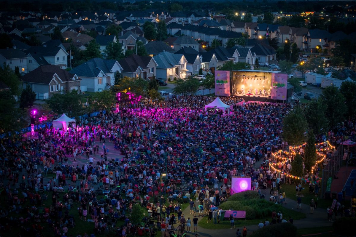 Vue aérienne d’un vaste spectacle extérieur en soirée, avec une foule dense rassemblée devant une scène illuminée au cœur d’un quartier résidentiel.