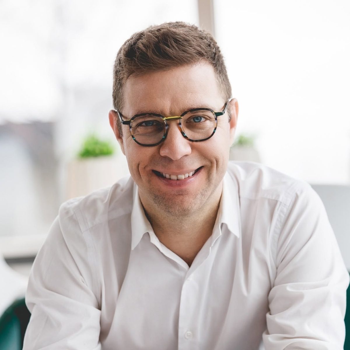 Une personne souriante portant des lunettes rondes, assise dans un espace lumineux avec une plante verte en arrière-plan flou