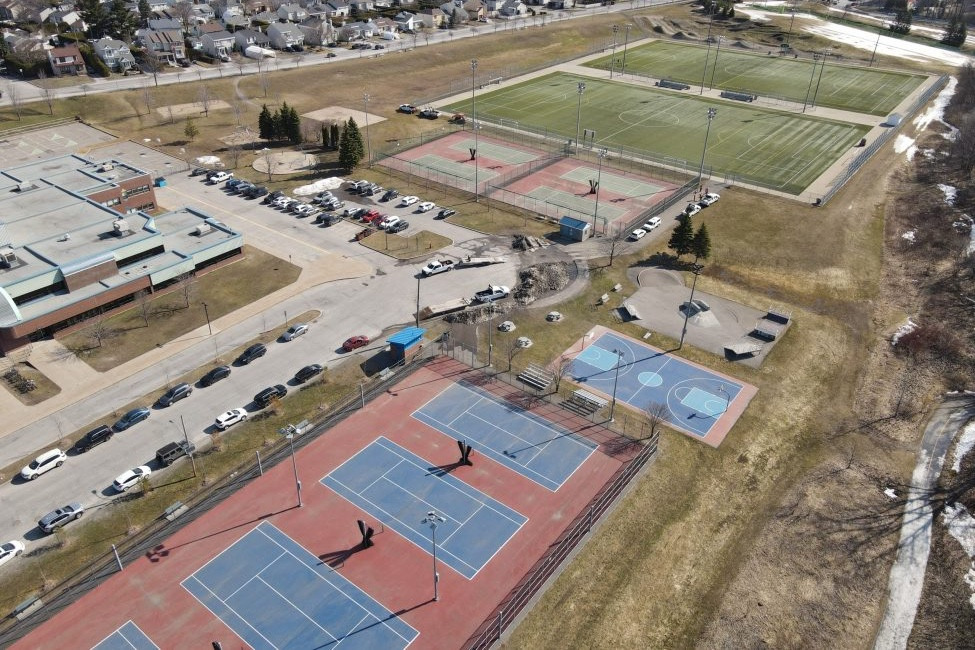 Vue aérienne d'un parc urbain avec terrains de tennis, basketball et soccer bordés de stationnements et de résidences