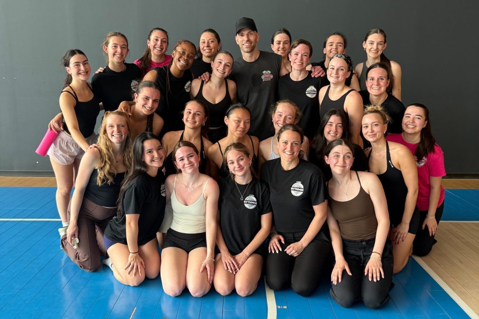 Groupe réuni dans un studio de danse ou d’entraînement. Les participants posent ensemble sur un plancher sportif bleu devant un mur sombre, dans une ambiance conviviale.