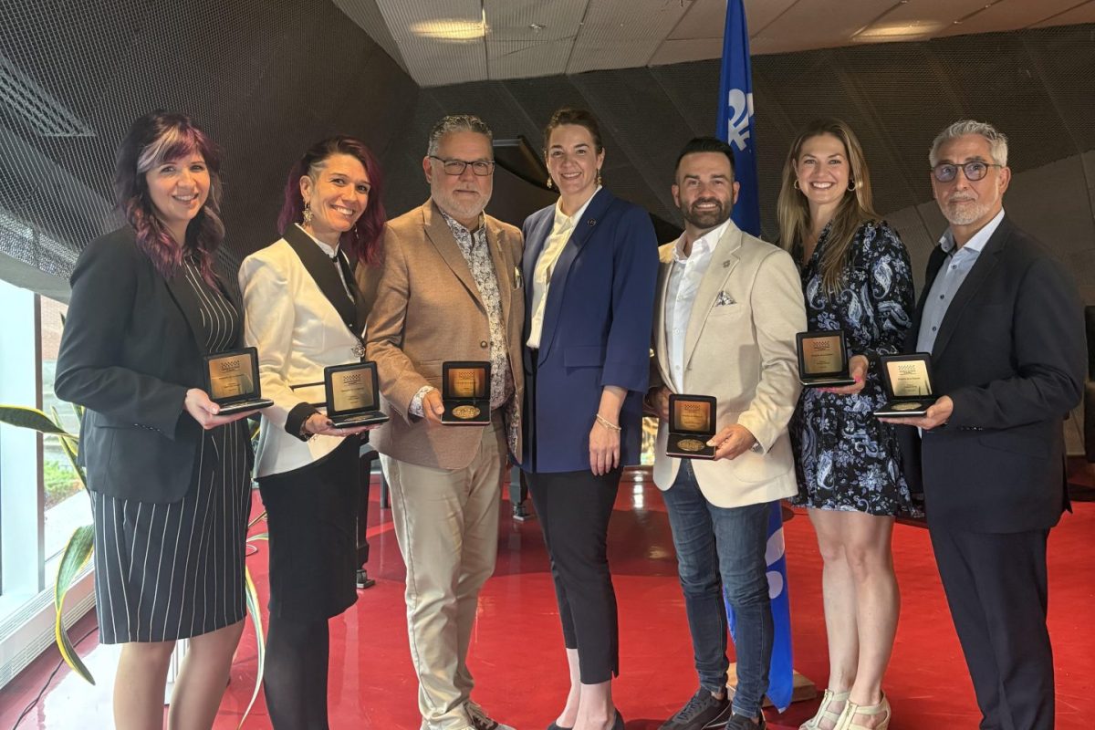 Groupe réuni dans un hall intérieur au sol rouge, tenant des plaques commémoratives devant un drapeau du Québec lors d’une cérémonie de reconnaissance.