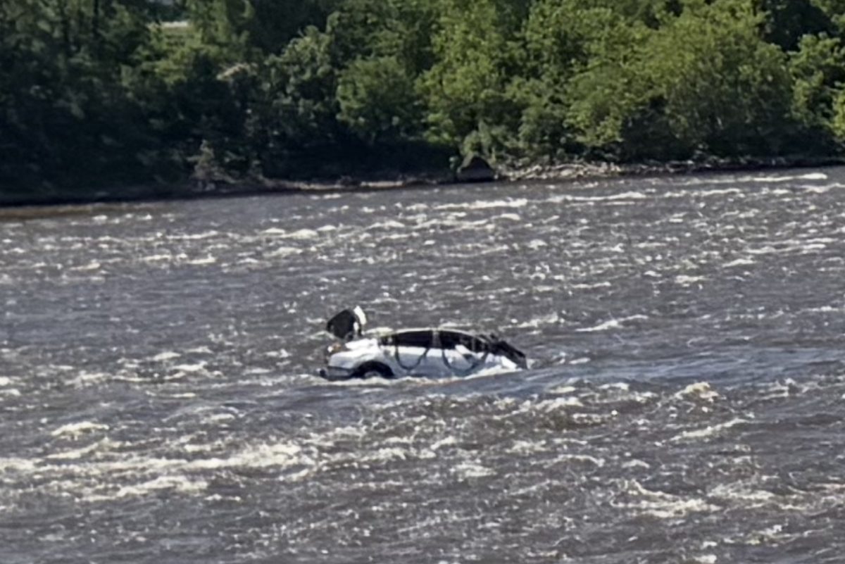 Une voiture presque entièrement submergée dérive dans un cours d'eau aux eaux turbulentes, avec une rive boisée en arrière-plan