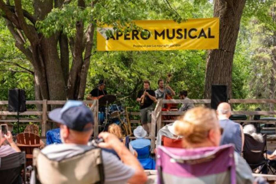 Groupe en concert en plein air sous bannière jaune « Apéro Musical », public assis en chaises de camping entouré d'arbres