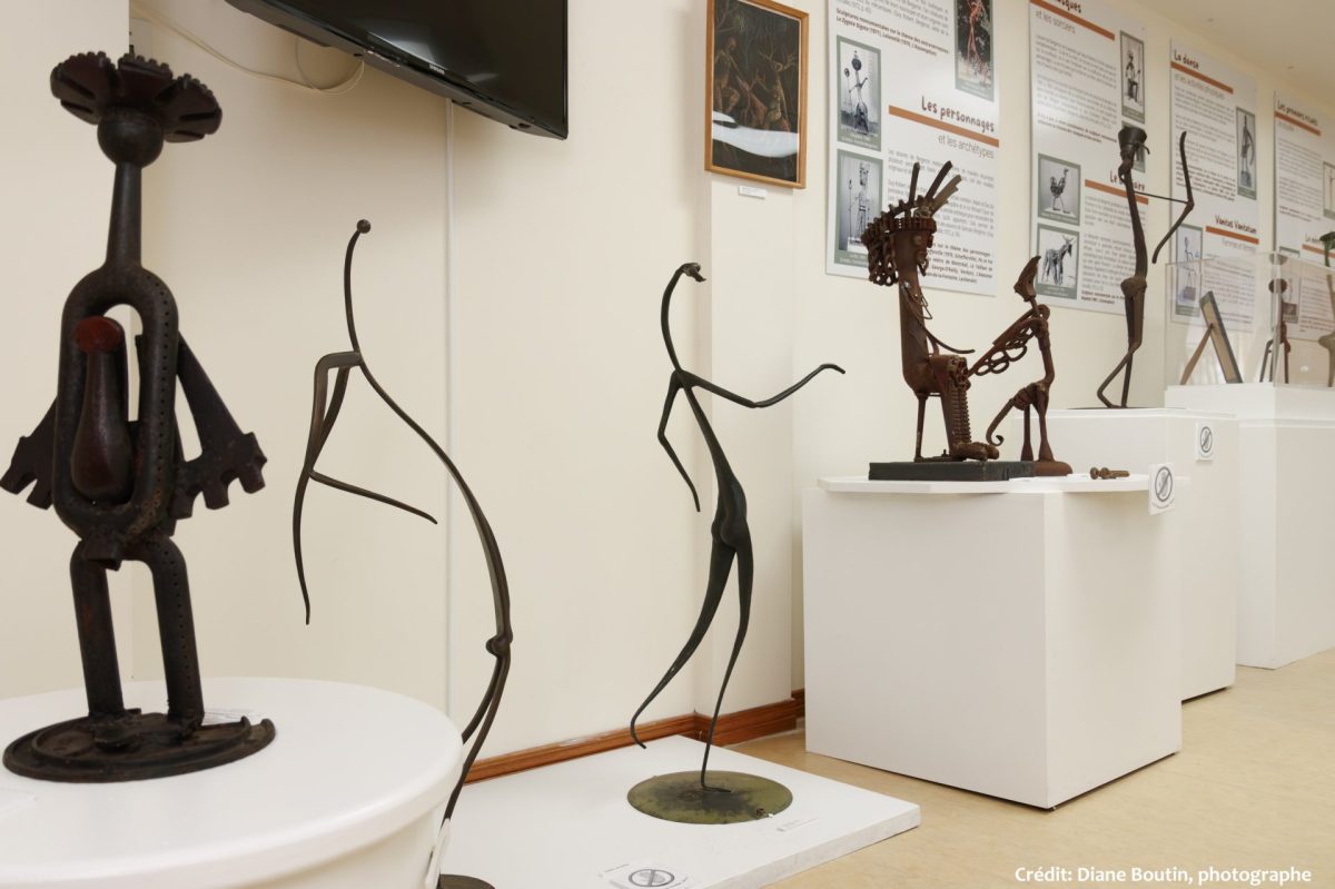 Salle d'exposition lumineuse avec sculptures métalliques figuratives sur socles blancs et panneaux explicatifs muraux
