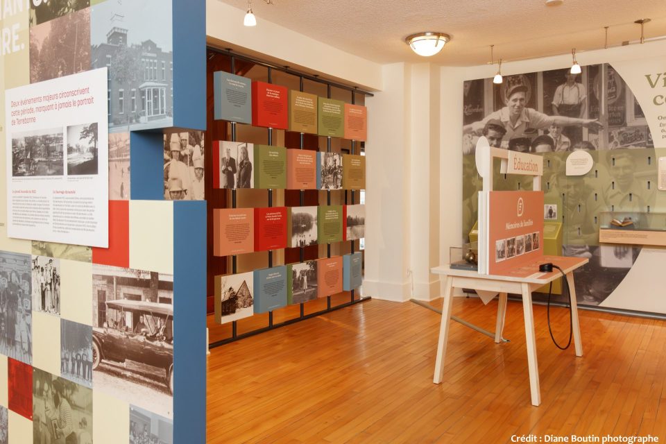Salle muséale avec panneaux colorés, photos historiques et une table interactive intitulée « Mémoires de familles »