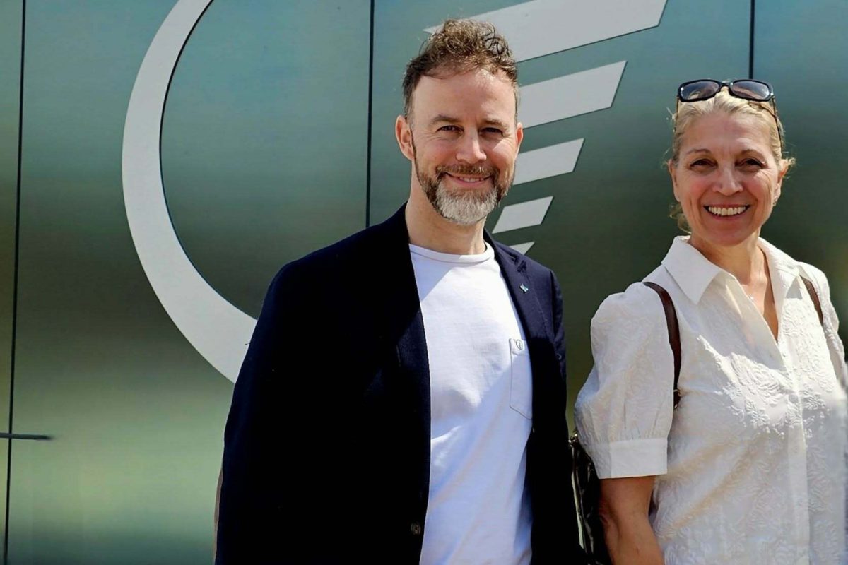 Deux personnes souriantes posent devant une façade métallique verte arborant un logo circulaire, par temps ensoleillé