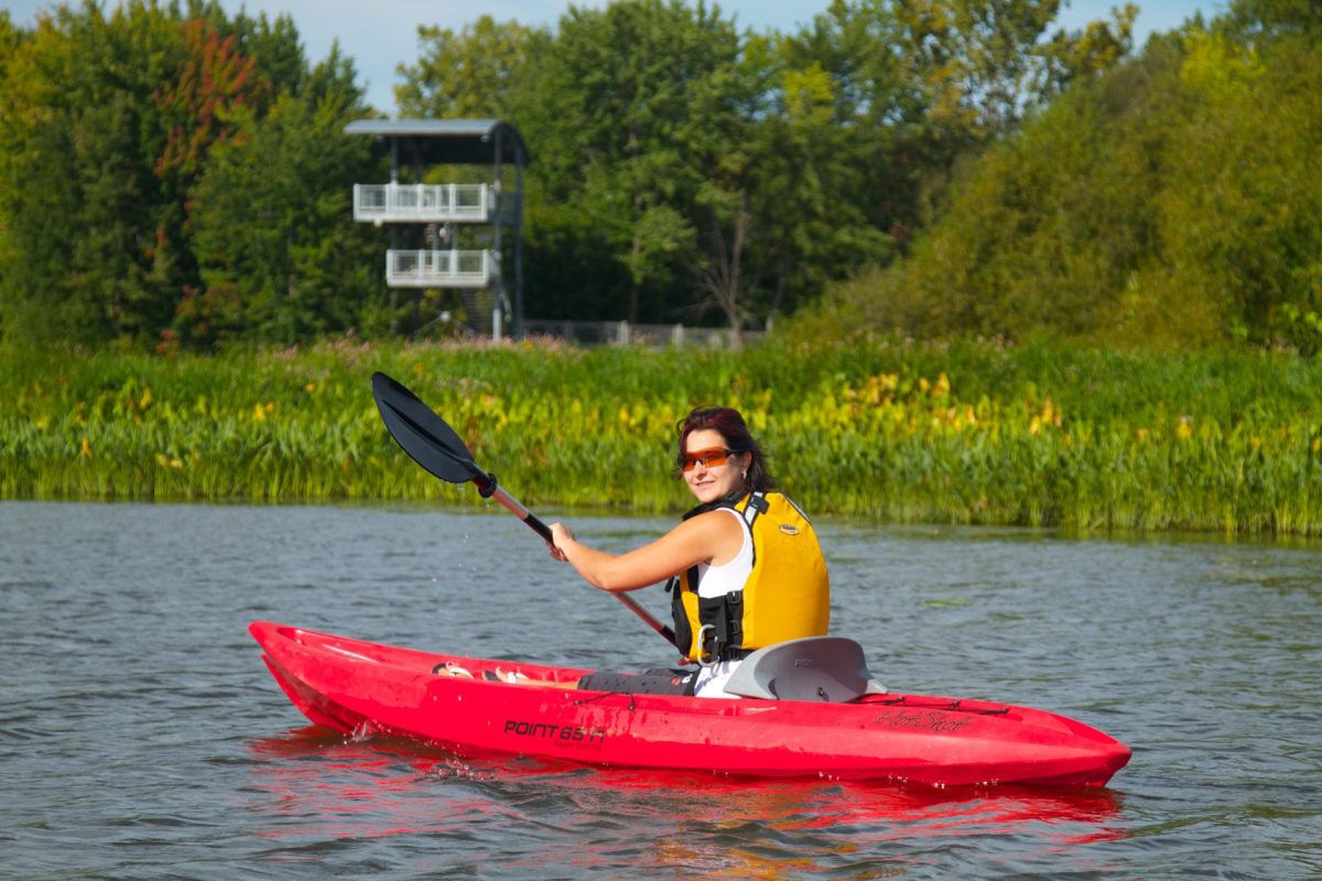 Kayak rouge naviguant sur une rivière bordée de végétation, avec une tour d’observation visible à l’arrière-plan sous un ciel dégagé.