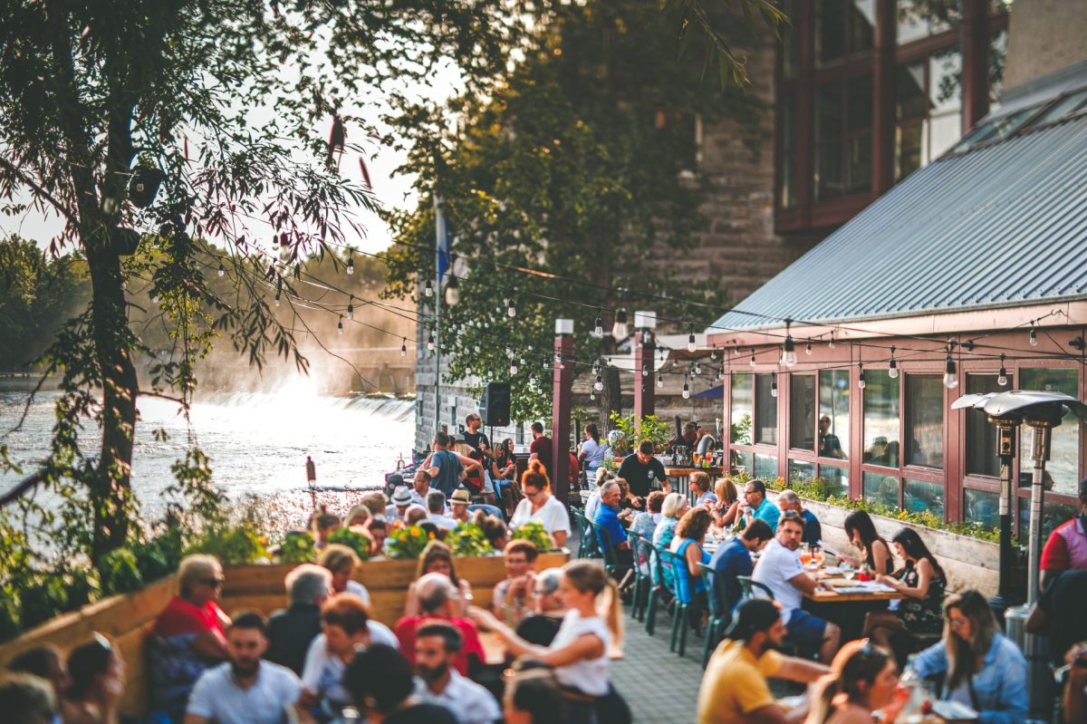 Terrasse en bord de rivière avec cascade, guirlandes lumineuses, personnes attablées, ambiance estivale