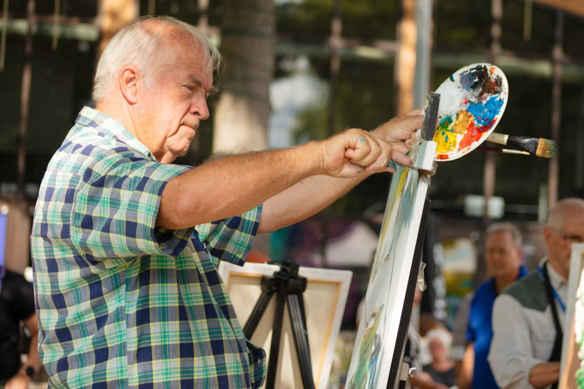 Un artiste peint sur une toile installée sur un chevalet extérieur, avec une palette colorée et d’autres participants en arrière-plan.