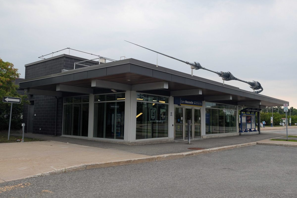 Bâtiment moderne de la Gare Mascouche avec façade vitrée, auvent plat et sculpture métallique sur le toit, sous un ciel nuageux