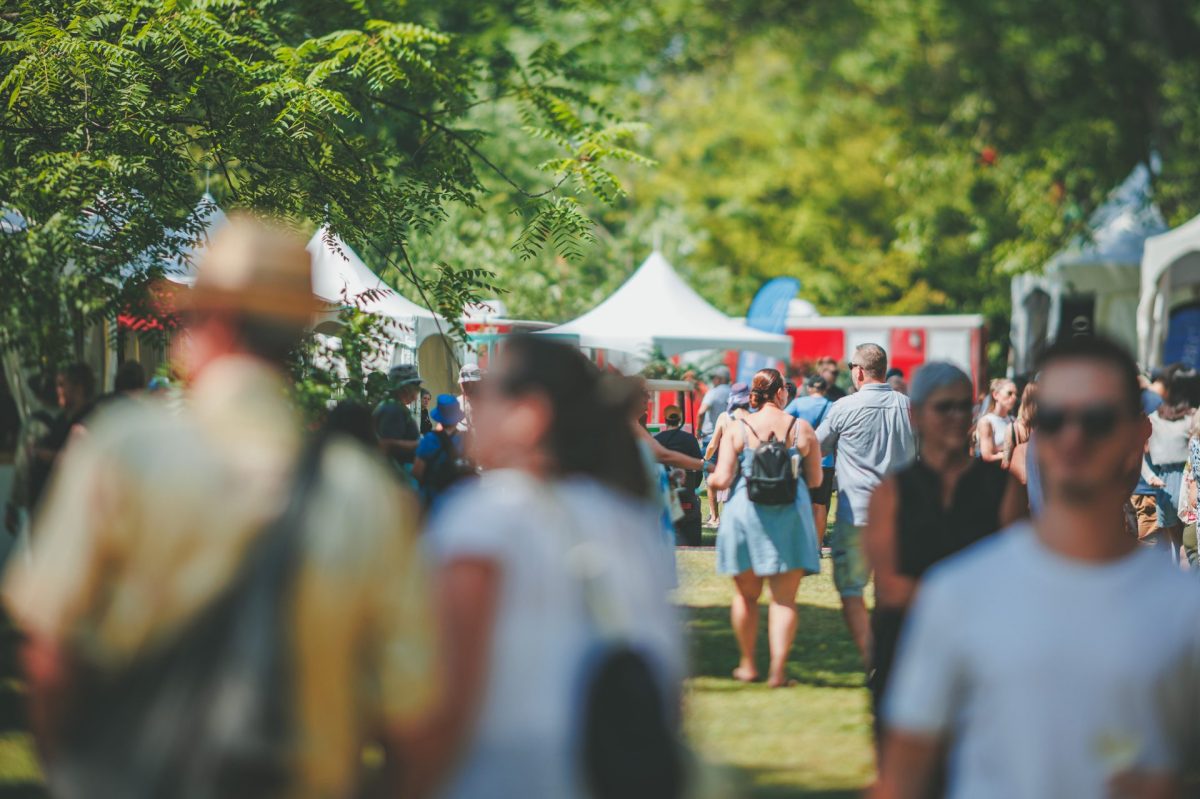 Une foule déambule dans un parc ensoleillé lors d'un festival estival, entre des chapiteaux blancs et des arbres verdoyants