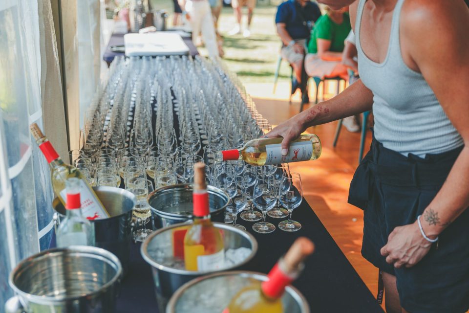 Stand de dégustation extérieur avec rangées de verres à vin, seaux réfrigérants et bouteille de blanc en cours de service