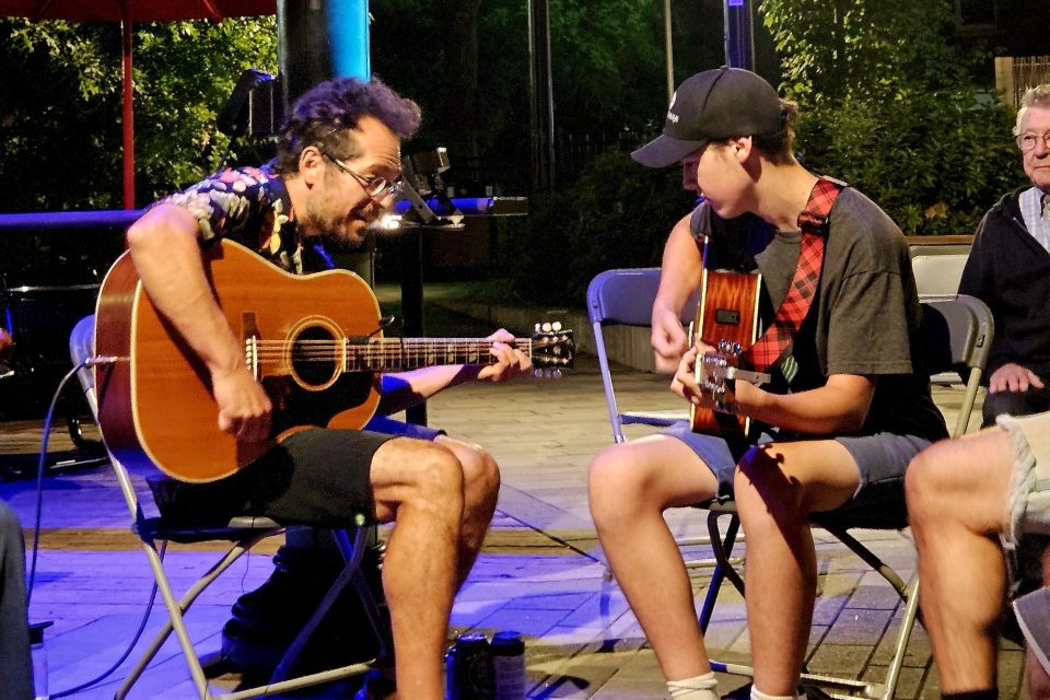 Deux personnes jouent de la guitare acoustique assises face à face en plein air de nuit, éclairées par une lumière violette