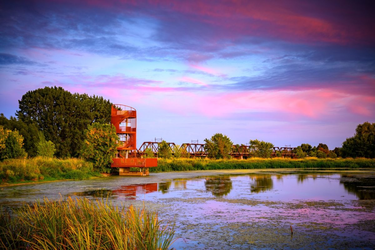 Tour métallique rouillée se reflétant dans un étang bordé de roseaux sous un ciel rose et violet au coucher du soleil