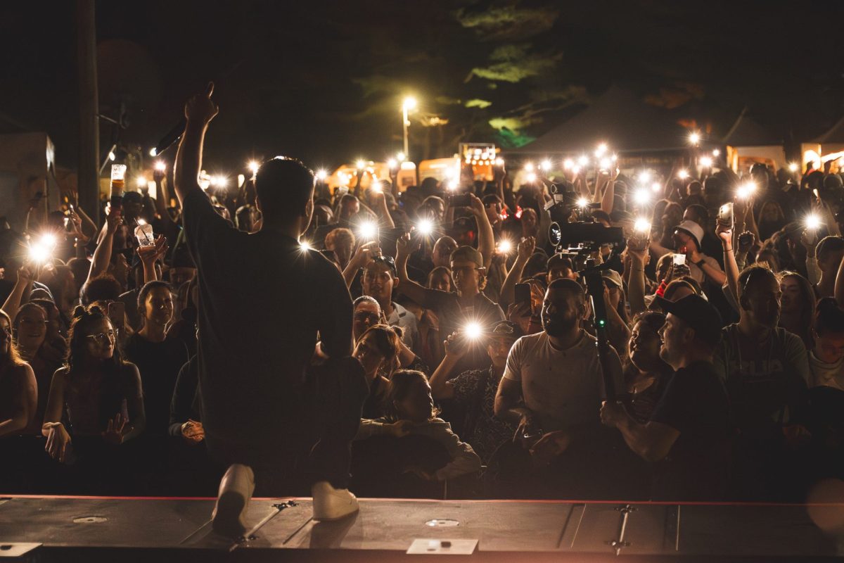 Sur scène en plein air la nuit, un artiste lève le bras devant une foule brandissant des lumières de smartphones