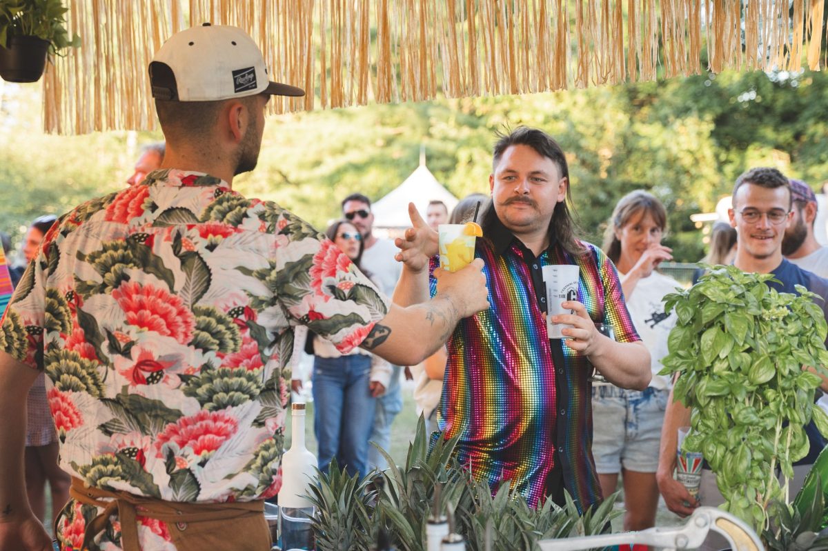Bar extérieur de festival estival, deux personnes s’échangent un cocktail garni d’orange parmi des plantes aromatiques