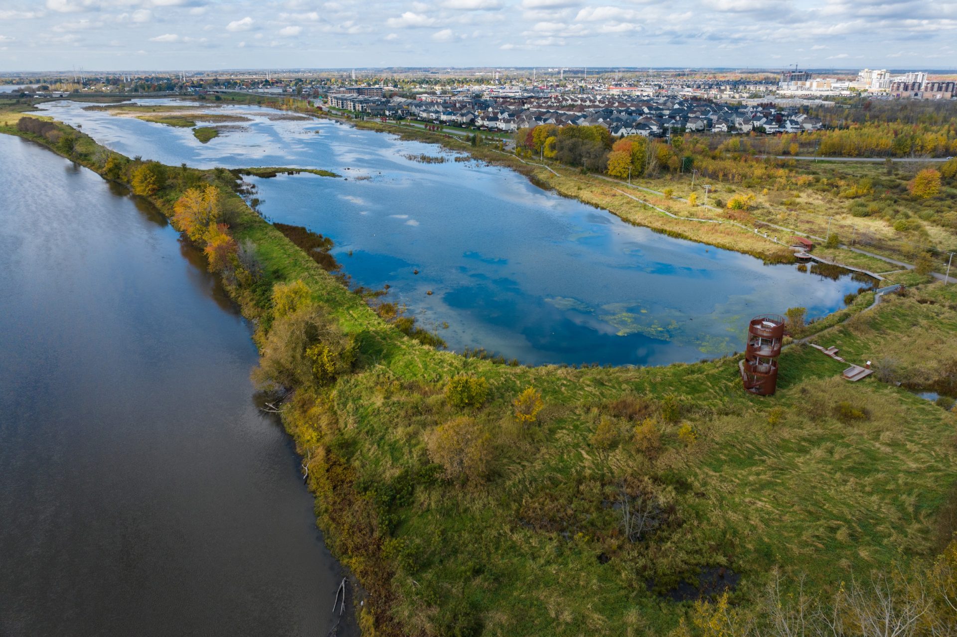 Vue aérienne d’une zone humide aux couleurs automnales, avec une tour d’observation en métal rouillé et une ville en arrière-plan