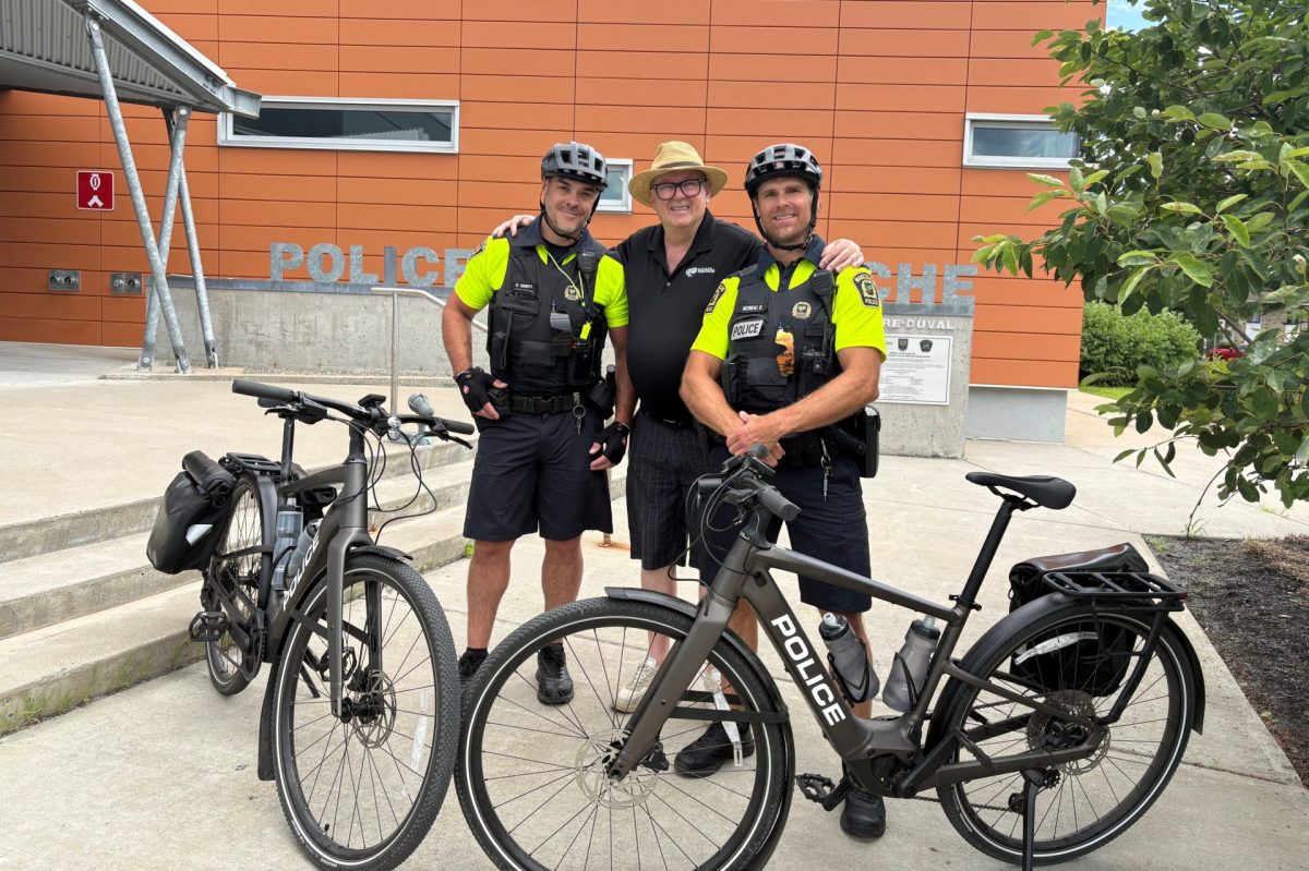 Trois personnes posent devant un poste de police avec deux vélos marqués « Police », par temps ensoleillé d'été