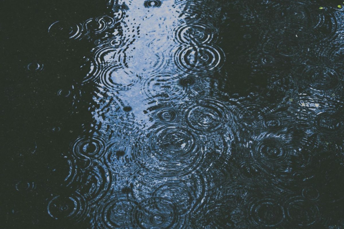 Des gouttes de pluie créent de nombreux cercles concentriques à la surface d’une eau sombre, où se reflète une bande de ciel bleu.